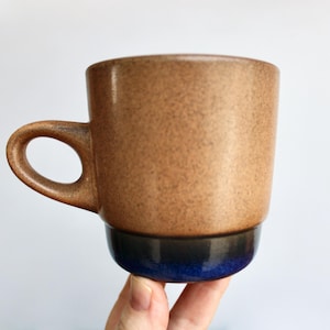 May include: A ceramic mug featuring a speckled light brown upper body and a smooth, dark blue glazed base. The mug has a rounded handle on the left side and a cylindrical shape with a slightly wider top rim, showcasing a two-tone color design.