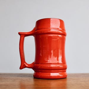 May include: A vibrant, glossy red ceramic beer stein with a large handle. The mug has a cylindrical body with a slightly flared base and a wide opening. The handle is curved and attached to the side of the mug.