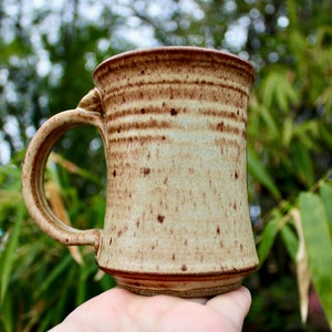 May include: A brown and tan ceramic mug with a speckled glaze. The mug has a wide, rounded base and a handle.