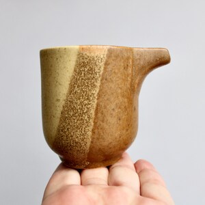 May include: A small, brown and beige ceramic creamer with a speckled glaze. The creamer has a rounded body and a spout.