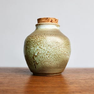 Może przedstawiać: Mały, ręcznie robiony ceramiczny słoik z korkiem. Słoik ma zaokrąglony korpus i teksturowane, nakrapiane szkliwo w odcieniach zieleni i brązu. Korek jest w kolorze jasnobrązowym.
