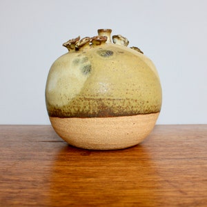 May include: A round, ceramic vase with a light brown glaze and a darker brown band around the middle. The vase has a unique, organic shape with small, rounded protrusions around the top.