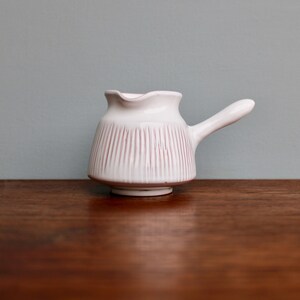 May include: White ceramic creamer with a pink striped design and a handle. The creamer is sitting on a wooden surface.