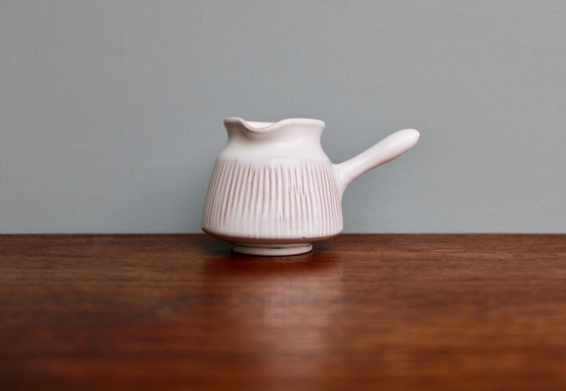 May include: White ceramic creamer with a pink striped design and a handle. The creamer is sitting on a wooden surface.