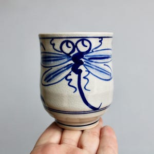 May include: A white ceramic mug with a blue dragonfly design. The dragonfly is painted in a stylized, abstract style.