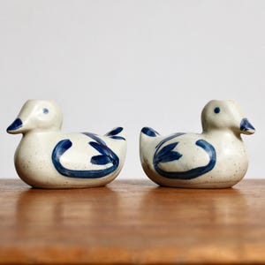 May include: Two ceramic duck figurines. The ducks are white with blue accents on the wings and body. The beaks are blue. The ducks are sitting on a wooden surface.