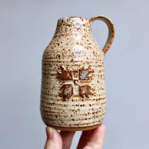 May include: A speckled ceramic pitcher with a handle. The beige-colored pitcher has a textured surface and a brown floral design. The pitcher is a decorative item.