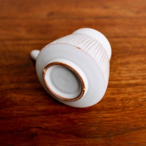 May include: White ceramic pitcher with a textured, striped design. The pitcher is shown from the bottom, revealing a brown rim.