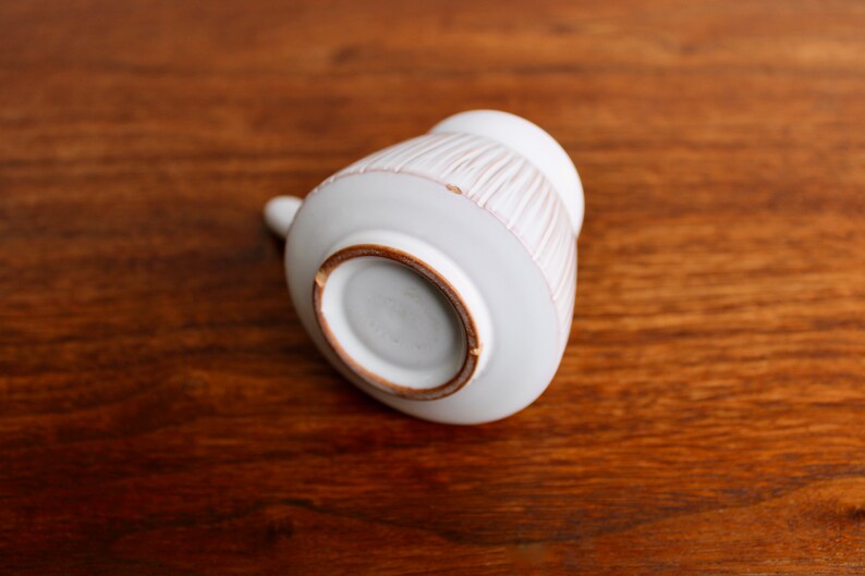 May include: White ceramic pitcher with a textured, striped design. The pitcher is shown from the bottom, revealing a brown rim.