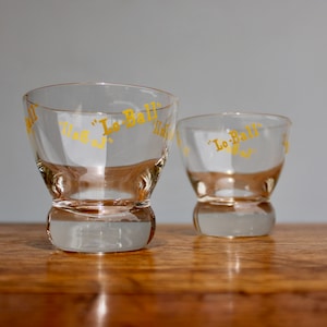 May include: Two clear glass shot glasses with a yellow "Lo-Ball" inscription. The glasses have a rounded bottom and a wide rim.