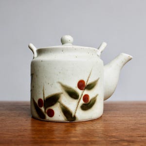 May include: A speckled, off-white ceramic teapot with a lid and a spout. The teapot is decorated with a pattern of green leaves and red berries. It has a handle on the lid and two small handles on the sides.