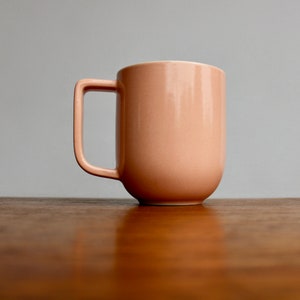 May include: A peach-colored ceramic mug with a square handle. The mug has a rounded base and a smooth, glossy finish. The mug is sitting on a wooden surface.