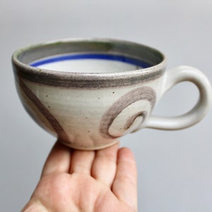 May include: A white ceramic mug with a brown and blue design. The mug has a wide, rounded shape and a handle.