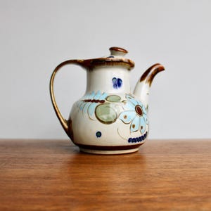 May include: A white ceramic teapot with a brown handle and spout. The teapot is decorated with blue, green, and brown floral designs.