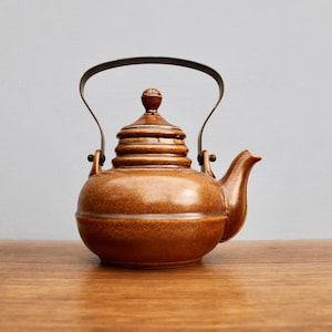 May include: A brown ceramic teapot with a metal handle and a layered lid. The teapot has a rounded body and a spout on the side.
