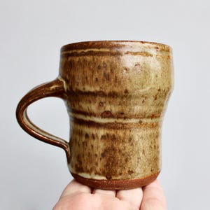 May include: A brown and tan ceramic mug with a handle. The mug has a rustic, handmade look.