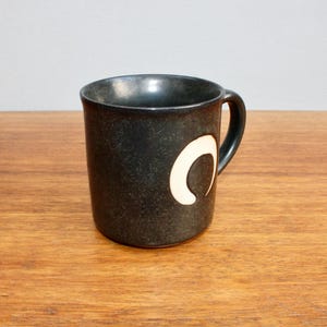 Pu&ograve; includere: Una tazza in ceramica grigio scuro con un design a mezzaluna bianca. La tazza ha un manico arrotondato.