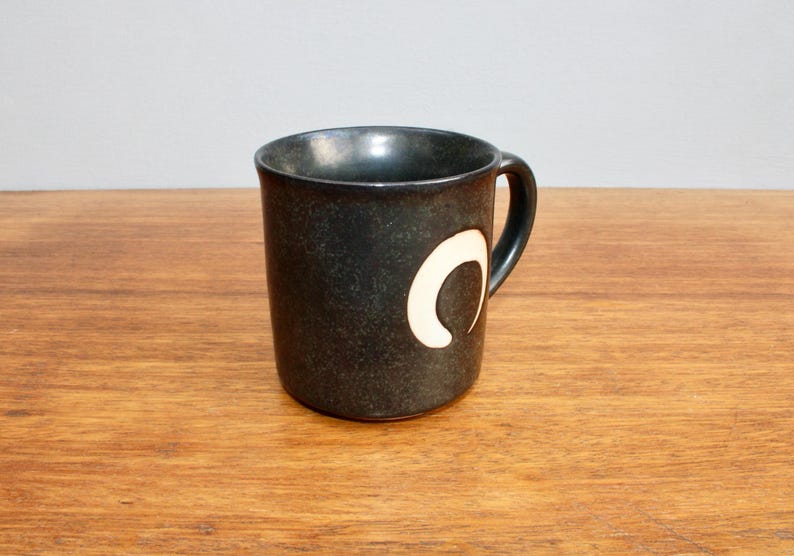 Pu&ograve; includere: Una tazza in ceramica grigio scuro con un design a mezzaluna bianca. La tazza ha un manico arrotondato.