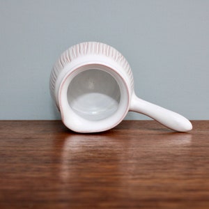 May include: White ceramic creamer with a ribbed design and a handle. The creamer is empty and sitting on a wooden surface.