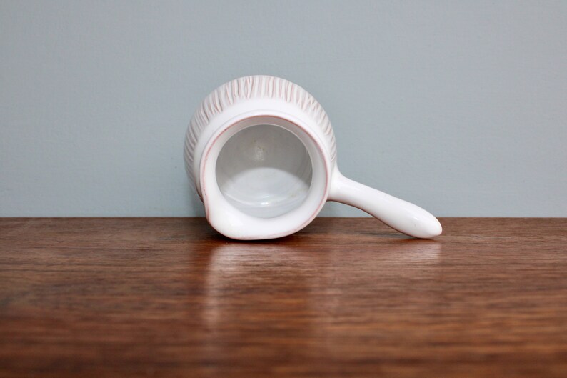 May include: White ceramic creamer with a ribbed design and a handle. The creamer is empty and sitting on a wooden surface.