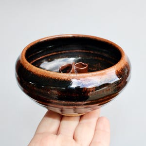 May include: A small, brown and black ceramic bowl with a unique design. The bowl has a slightly flared rim and a smooth, glossy finish.