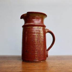 May include: A red and brown ceramic pitcher with a handle. The pitcher has a rustic, handmade look.