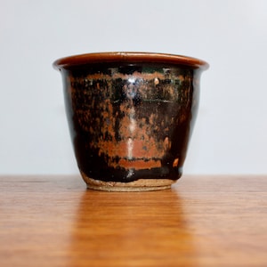 May include: A small, handcrafted ceramic pot with a dark brown exterior and a lighter brown interior. The pot has a slightly flared rim and a textured surface, perfect for holding small plants or decorative items.
