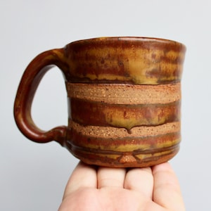 May include: A brown ceramic mug with a striped design. The mug has a wide, rounded handle and a slightly flared rim.