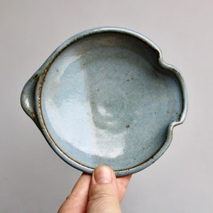 May include: A light blue ceramic dish with a scalloped edge and two handles. The dish has a rustic, handmade look.