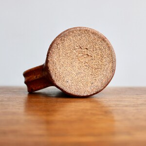 May include: A brown ceramic mug with a textured surface. The bottom of the mug has a brown glaze with a stamped mark that reads "The Pottery Place".
