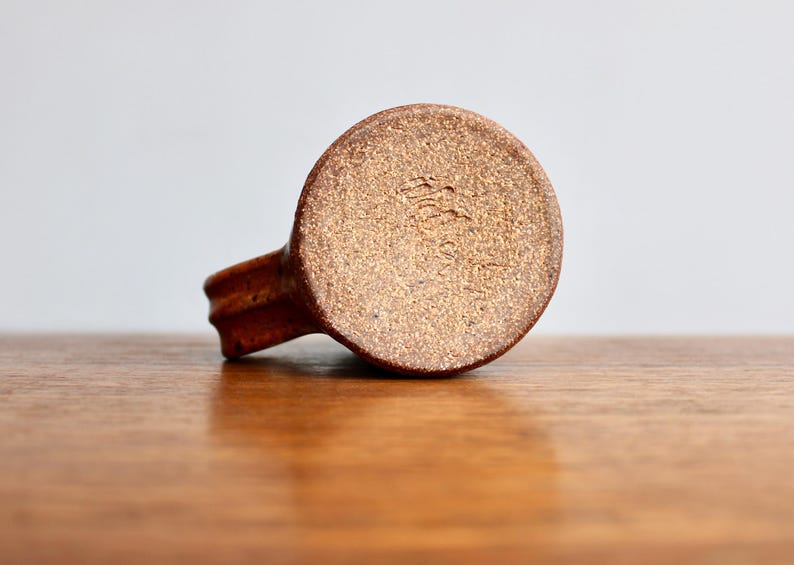 May include: A brown ceramic mug with a textured surface. The bottom of the mug has a brown glaze with a stamped mark that reads "The Pottery Place".
