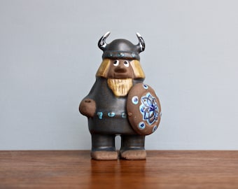 May include: A ceramic figurine of a Viking warrior with a horned helmet, holding a shield with a blue and white floral design. The figurine is painted in shades of brown, blue, and white.