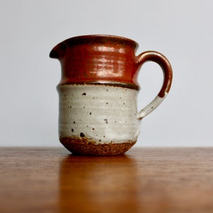 May include: A small, brown and white ceramic pitcher with a handle. The pitcher has a speckled glaze and a rounded base.