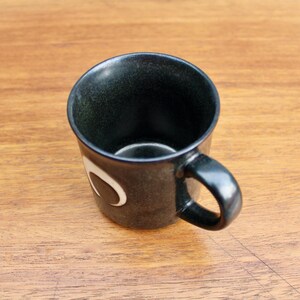 Pu&ograve; includere: Una tazza in ceramica nera con un design a mezzaluna bianca. La tazza ha un manico arrotondato.