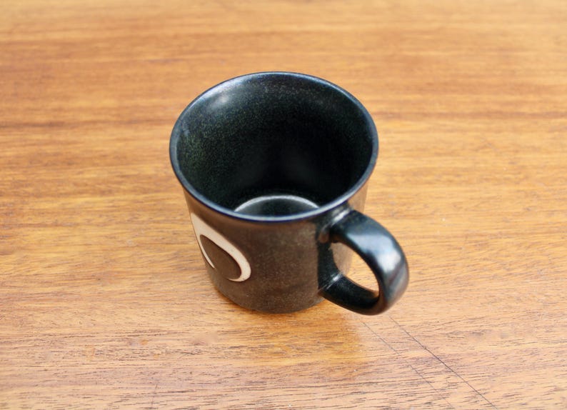 Pu&ograve; includere: Una tazza in ceramica nera con un design a mezzaluna bianca. La tazza ha un manico arrotondato.