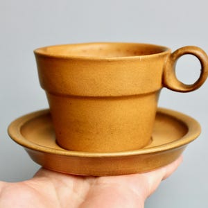 May include: A brown ceramic coffee cup and saucer set. The cup has a unique, stacked design and a rounded handle. The saucer is a simple, round shape.