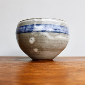 May include: A small, round, gray ceramic bowl with a blue and white stripe around the middle. The bowl has a slightly textured surface.