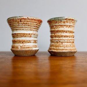 May include: Two handmade ceramic vases with a unique design. The vases are white with horizontal brown stripes and a speckled texture. The bases are a darker brown. The rims have a green glaze. The vases are displayed on a wooden surface.