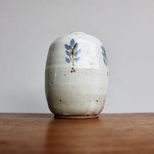 May include: A white ceramic vase with a blue and brown floral design. The vase has a rounded shape and a slightly textured surface.