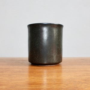 Pu&ograve; includere: Un vaso in ceramica verde scuro con una finitura macchiettata. Il vaso ha una forma cilindrica e un bordo liscio e arrotondato.