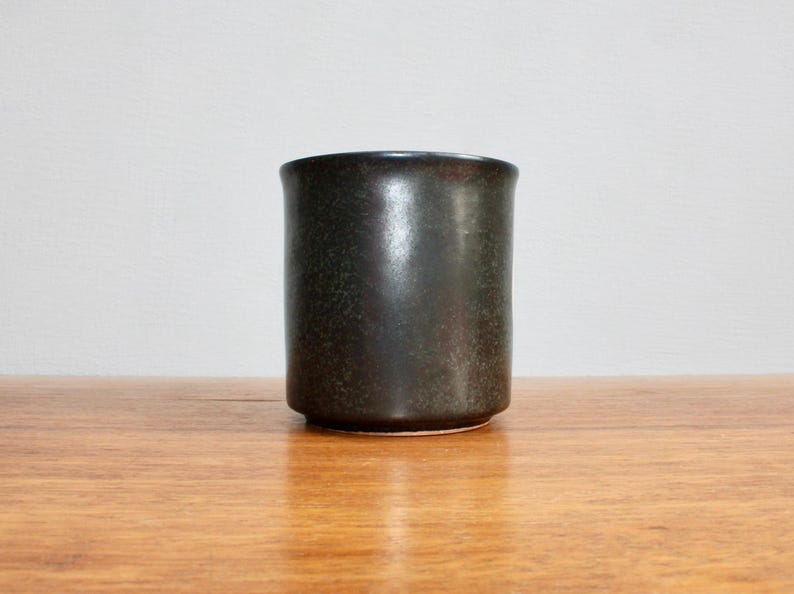 Pu&ograve; includere: Un vaso in ceramica verde scuro con una finitura macchiettata. Il vaso ha una forma cilindrica e un bordo liscio e arrotondato.