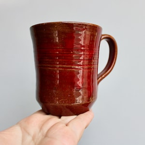 May include: A red ceramic mug with a rounded shape and a handle. The mug has a glossy finish and a subtle brown glaze.