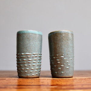 May include: Two teal ceramic shot glasses with a textured pattern. The glasses are on a wooden surface.