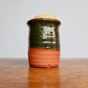 May include: A handmade ceramic jar with a cork stopper. The jar is cylindrical, with a dark green upper section, a reddish-orange middle, and a cork stopper. The jar sits on a wooden surface.