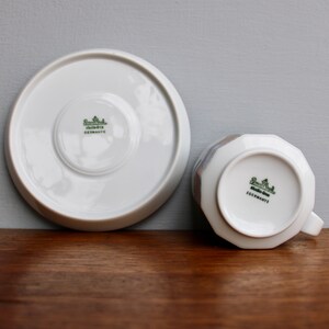 Może przedstawiać: Biały zestaw filiżanki i spodka z porcelany. Filiżanka ma fasetowany wz&oacute;r i uchwyt. Spodek i dno filiżanki mają zielone logo i napis "GERMANY". Zestaw znajduje się na drewnianej powierzchni.