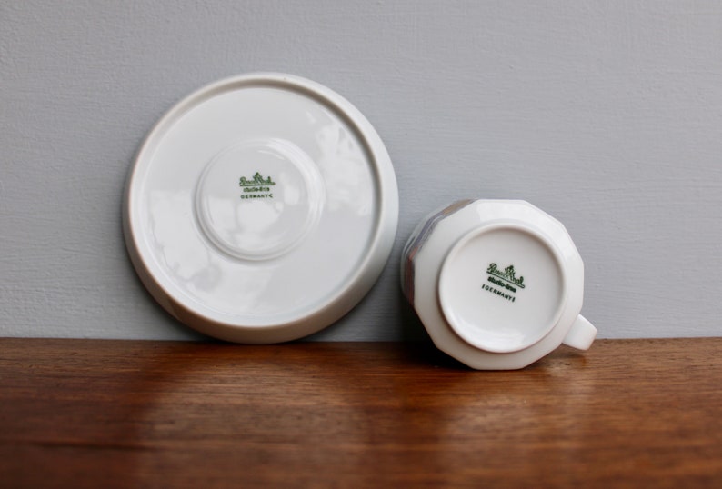 Może przedstawiać: Biały zestaw filiżanki i spodka z porcelany. Filiżanka ma fasetowany wz&oacute;r i uchwyt. Spodek i dno filiżanki mają zielone logo i napis "GERMANY". Zestaw znajduje się na drewnianej powierzchni.