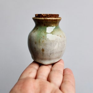 May include: A small, white ceramic jar with a green and brown glaze. The jar has a cork lid and is sitting on a hand.