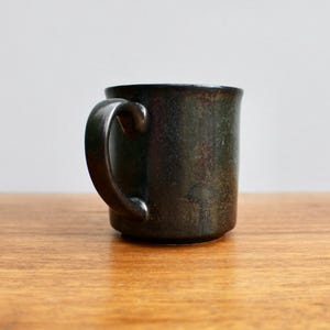 Pu&ograve; includere: Una tazza in ceramica grigio scuro con una glassa macchiata. La tazza ha un manico arrotondato.