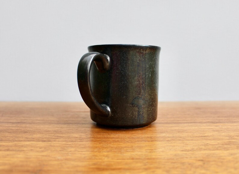 Pu&ograve; includere: Una tazza in ceramica grigio scuro con una glassa macchiata. La tazza ha un manico arrotondato.