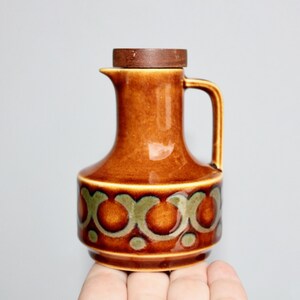May include: A vintage brown ceramic pitcher with a wooden lid. The pitcher has a handle and is decorated with a repeating pattern of brown and green circles. The pitcher is held by a hand.